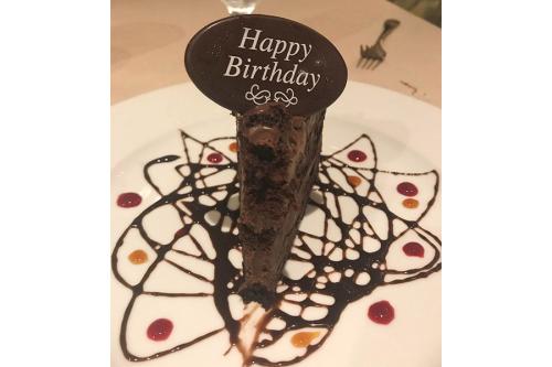 A small slice of chocolate cake on a white plate drizzles with chocolate syrup; the cake slice is topped with a chocolate decorative oval that says Happy Birthday. 
