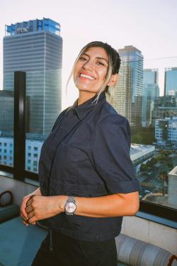 A woman in a black chef's coat smiles on rooftop balcony, skyscrapers in the distance.