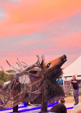 Under a bright pink sky, a figure of a giant elk head with long, branched antler lifts its chin. It is supported by two people underneath a another person holding draped fabric to the side.