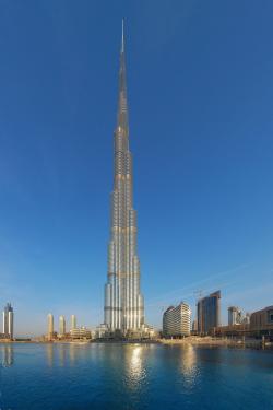 Spire-like skyscaper against blue sky and reflected in blue water in the foreground.