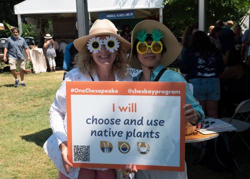 Two women wearing tan sun hats and novelty glasses, one with white flowers as lenses and the other as pineapples, hold up a sign that reads: I will choose and use native plants.