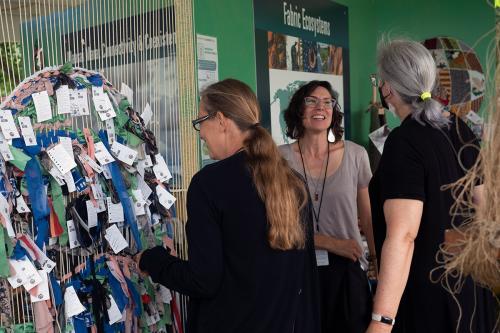 Three people stand under a sign that reads Fabric Ecosystems. To their left, a large makeshift loom of vertical white strings contains a circular pattern of fabric scraps in green, blue, pink, and black.