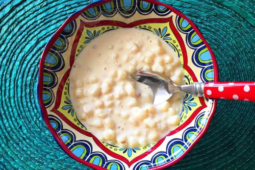 A bowl of creamy white pudding in a colorful patterned bowl, with a bright red spoon sticking out and bright turquoise placemat underneath.