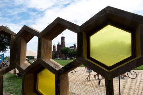 A large wooden honeycomb structure with yellow translucent panels frames the red-brick Smithsonian Castle.