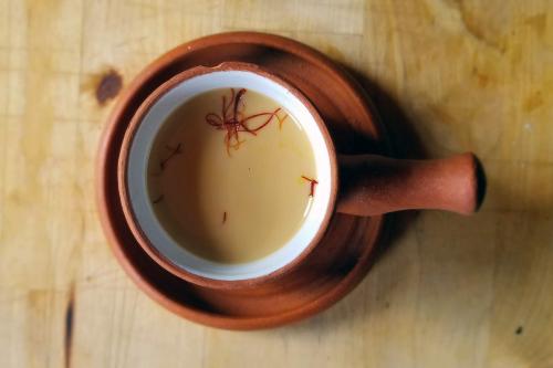 Reddish borwn mug full of milky tan chai, with bright red threads of saffron floating on top