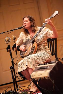 A woman in a white patterned dress with wavy dirty blond hair plays banjo on stage.