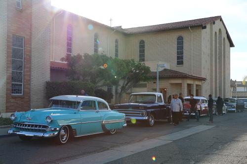 Several classic cars parked in front of a church building, including a shiny turquoise car with chrome details. A small crowd of people stands in the street.