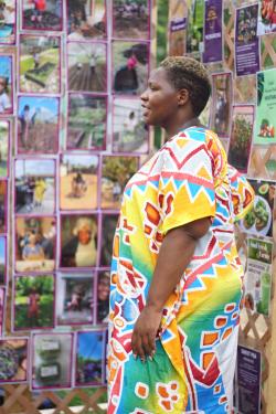 A woman stands in a bright, multicolored dress, speaking to visitors out of frame. Behind her is a wall with a grid of photos of different people, farms, and plants.