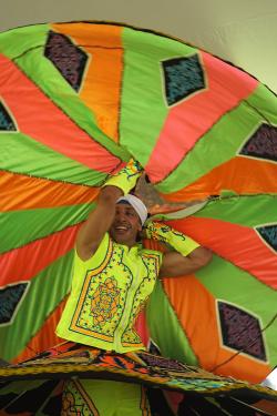 A man wearing bright colors twirls a large piece of circular fabric over his head, his skirt twirling alongside of it.