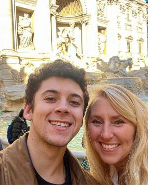 A woman and young man pose together and smile while taking a selfie in front of the Trevi Fountain in Rome, Italy.