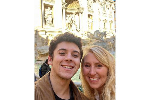 A woman and young man pose together and smile while taking a selfie in front of the Trevi Fountain in Rome, Italy.