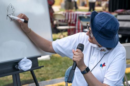 A woman, seated and holding a microphone, draws a heart-shaped bird on the white board with a marker. 