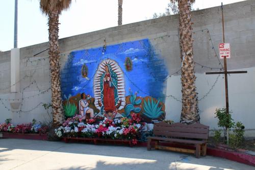 Painting of the virgin on a cement brick wall between two palm tree trunks, with a No Parking sign and a brown bench to the side. At the base of the mural, several bouquets of white and red flowers.