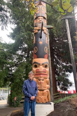 An elder man poses in front of a completed totem pole outdoors, with figures carved and painted in black and red.