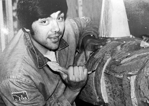 A young man smiles at the camera as he carves into a large piece of wood with a hand tool. On his denim jacket is a patch with the Alaskan state flag, a constellation of stars. Black-and-white photo.