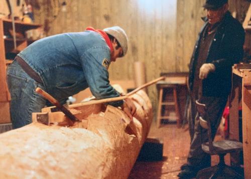A young man works on carving a tree trunk laid horizontally in a workshop while an older man oversees. Old color photo.