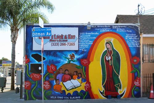 Mural on the side of a corner commercial building, featuring a virgin figure, red roses, a group of kids around a book, and white text with artists' names. A street sign in front reads Ditman Ave.