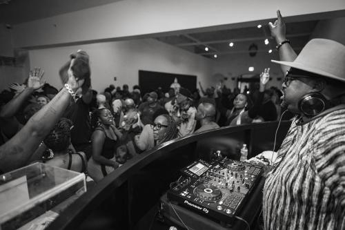 A man in a DJ booth raises one hand in front of a crowd of dancers.