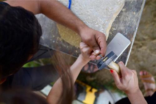 Seen from above, two sets of hands fiddle with stone carving tools leaning on a work table with a stone carving slab. The hair of the person on the left falls into the frame.
