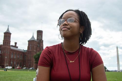 A young woman with short black dreads and a glucose monitor on her upper arm looks into the distance, sitting in the grass of the National Mall, with the Smithsonian Castle and Washington Monument in the background.