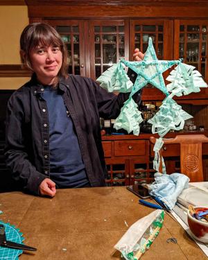 A woman holds up a bright blue star made out of tissue paper, with ornaments hanging from the four bottom rays.
