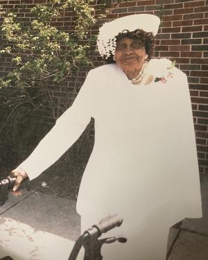 An elder Black woman in all-white church outfit poses in front of a brick wall. She holds onto a walker.