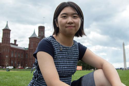 A young woman with short black hair sits in the grass of the National Mall, with the Smithsonian Castle and Washington Monument in the background.