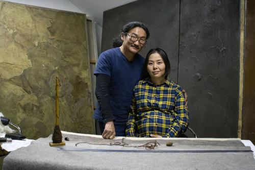 A man, standing, poses with a seated, pregnant woman, amid gray and brown textiles on the table in front of them and walls behind them.