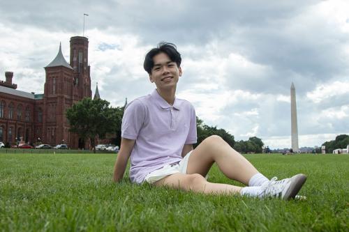 A young man with short black hair sits in the grass of the National Mall, with the Smithsonian Castle and Washington Monument in the background.
