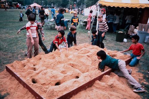 Six kids play in and around a rectangular sandbox set in the grass among festival tents. Someone has burrowed gopher-like holes into the sand. Old color photo.
