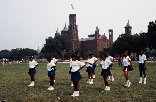 Eleven young women in matching white T-shirts, blue miniskirts, and white tennis shoes stand in formation on the green grass of the National Mall, with the red-brick Smithsonian Castle behind them.