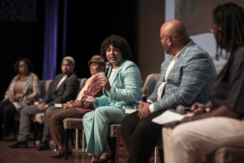 From the side, six people sit lined up on a stage. A woman in a turquoise seat faces her fellow panel members, speaking into a microphone.