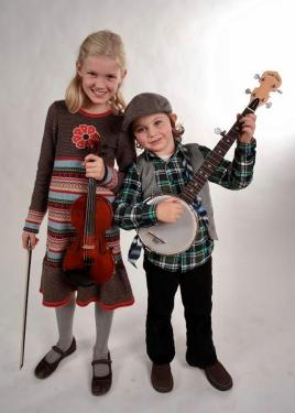 A young girl and boy pose for a studio photo together; the girl holds a fiddle and the boy a banjo.
