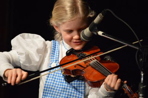 A young girl in a white blouse and blue gingham pinafore smiles as she plays fiddle in front of a microphone.
