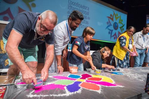 Indoors, at the front of a conference hall, adults and a child work on a multicolored flower carpet made of sawdust on a low platform.