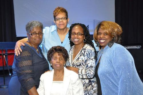 Five Black women dressed in shades of blue and white pose together, smiling.