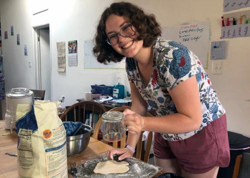 A young woman leans over a table, forming a piece of dough on a cutting board. She looks up at the camera, smiling.