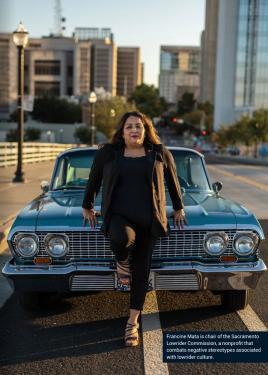 A woman poses leaning against the hood of a lowrider car in a city street.