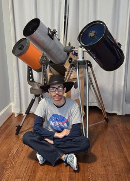 A young man with a black fedora and NASA T-shirt sits cross-legged on a floor in front of three cyclindral telescopes on tripods.