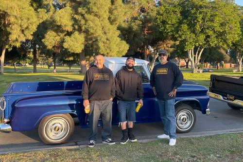 Three men pose in a park, standing in front of a royal blue classic truck.