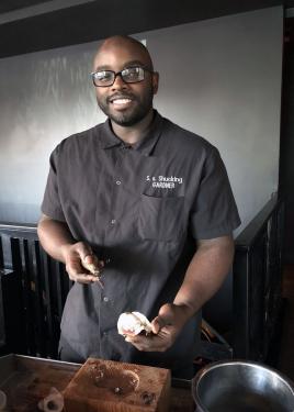 A Black man in black chef's coat holds an oyster in one hand and shucking knife in another.