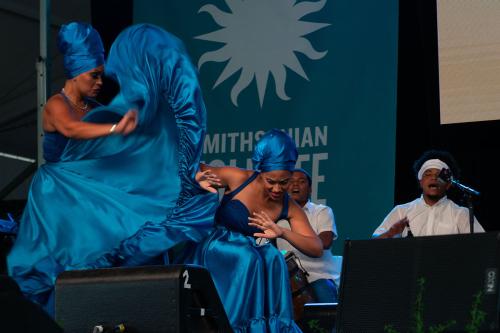 Nine singers in traditional Ukrainian dress sing into microphones on the Ralph Rinzler Main Stage, with a video of a man playing a traditional Ukrainian plucked string instrument.