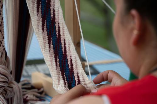 A woman threads a multicolored thin tapestry across a loom, with pink, dark red, blue, and beige zigzags. 