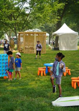 A young child stands in a field of grass with a small bean bag in his hand, preparing to throw it on a cornhole board. Behind him is a large connect four set.
