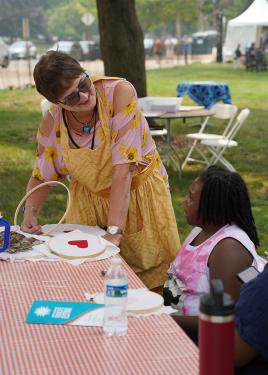 A young girl sits at a table with a red and white checkered tablecloth. She looks up at an older woman who holds an embroidery hoop. On the table are supplies for embroidery and other craft projects.