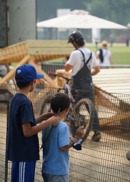 Two young boys stand at a fence looking out over it. In their view, a mountain biker prepares for a ride down the Festival’s custom wooden track.