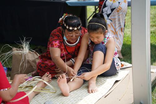 A woman instructs a child how to braid plant fiber.
