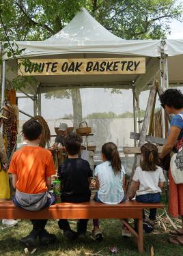 Four kids sit on a bench facing the White Oak Basketry tent, watching a man demonstrate wood shaving.