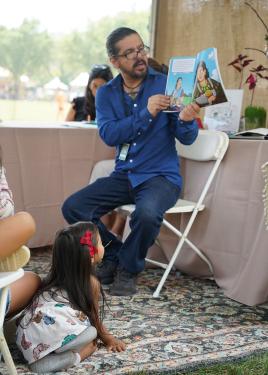 A little girl sits on a carpet looking up at a man who is reading stories from a children’s book.