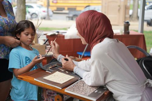 A woman leans over a calligraphy desk and touches a child’s finger with her quill.